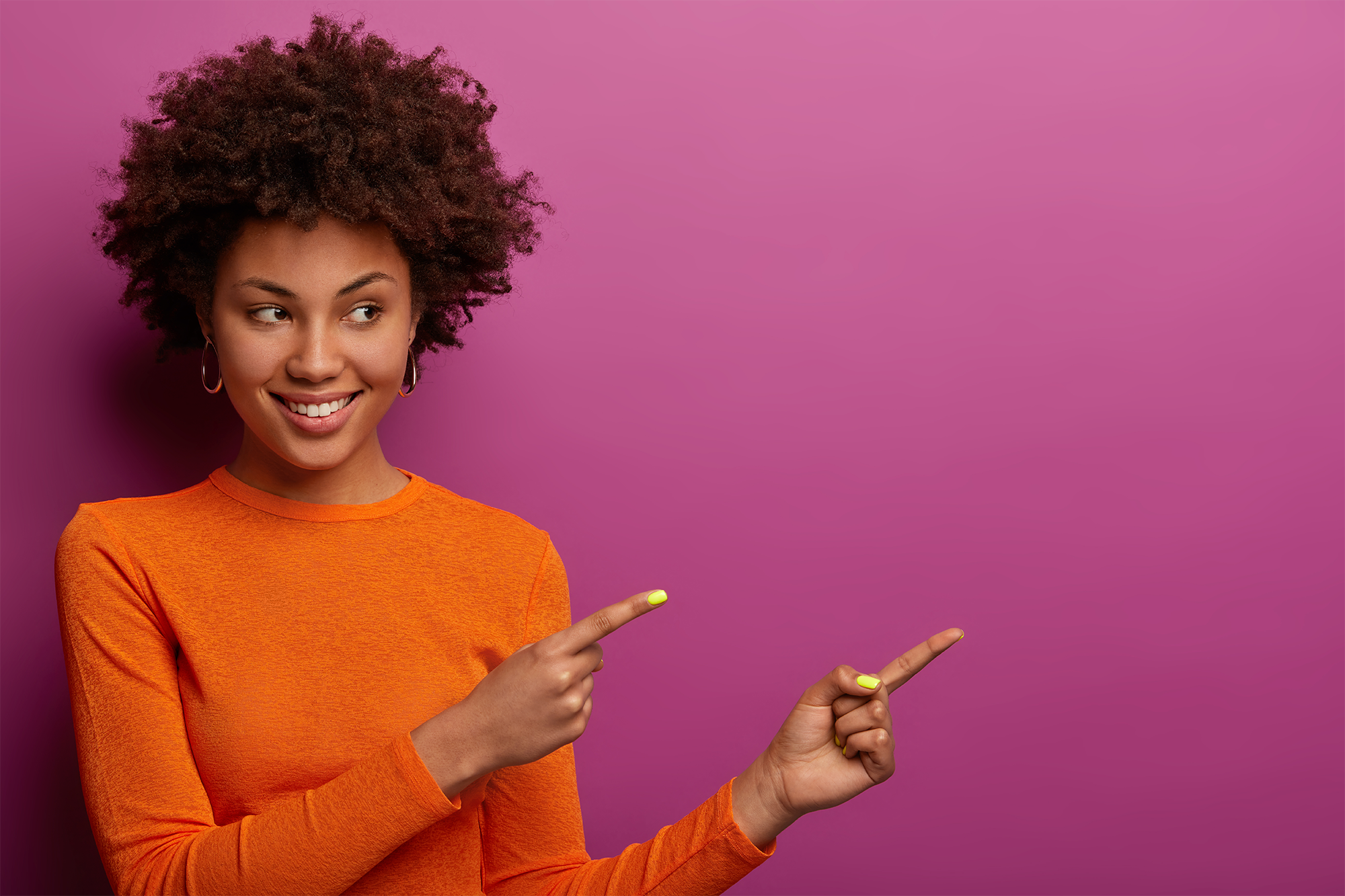 Femme avec des cheveux frisés qui pointe ses doigts vers la droite. Il y a un fond rose derrière.